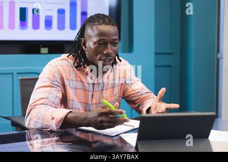 Homme afro-américain assis à la table de conférence, tenant un stylo vert et faisant des gestes vers la tablette Banque D'Images