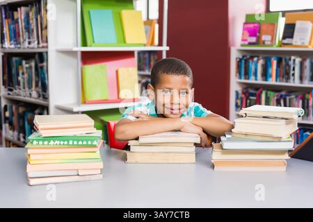 Garçon appuyé sur trois piles de livres dans la bibliothèque de l'école, avec une table grise et des classeurs colorés Banque D'Images