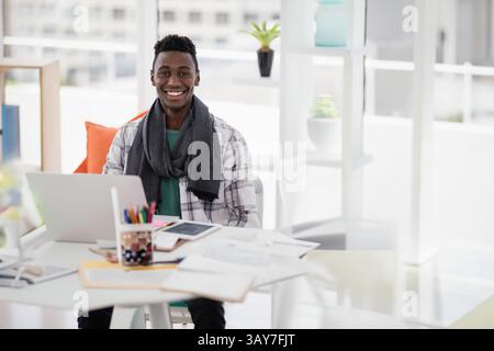 Jeune homme afro-américain utilisant un ordinateur portable, tablette au bureau dans l'espace de travail avec des papiers, des crayons de couleur Banque D'Images