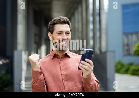 Un homme dans une chemise rouge rayonne de joie alors qu'il vérifie son téléphone, serrant son poing dans l'excitation à l'extérieur. Banque D'Images