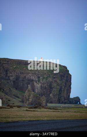 Phare au sommet d'une falaise spectaculaire près de Dyrhólaey, Islande, sous un ciel bleu clair au crépuscule, avec un premier plan herbeux et des formations rocheuses volcaniques. Banque D'Images