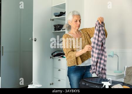 Femme senior triant les vêtements de la valise noire dans la chambre minimaliste, avec armoire intégrée Banque D'Images