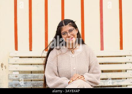 Une jeune femme pose sur un banc, mettant en valeur ses tricots élégants et son sourire joyeux contre des rayures vibrantes. Banque D'Images