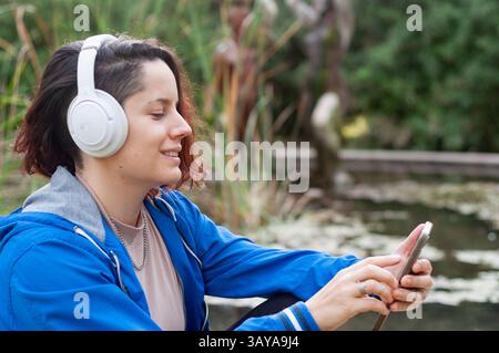 Heureuse jeune femme portant des écouteurs sans fil choisit la musique de playlist sur son application smartphone, assise dans un parc avec étang et arbres à l'arrière Banque D'Images