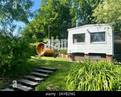 Une scène arrière unique et confortable avec une maison de jardin blanche moderne et un sauna en baril élégant. Entouré d'une verdure vibrante et ombragé par de hauts t Banque D'Images