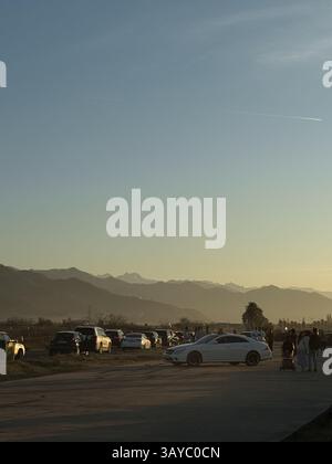 Cette image montre une scène de bord de route pendant le coucher du soleil. Plusieurs voitures et personnes stationnées, y compris un groupe avec une poussette de bébé, sont visibles au premier plan Banque D'Images