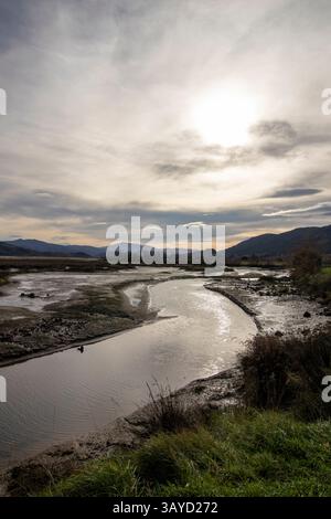 Une rivière sinueuse coule à travers un paysage boueux sous un ciel nuageux avec le soleil partiellement caché, créant une scène sereine Banque D'Images