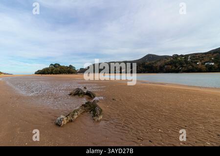 Un rivage de sable paisible rencontre l'eau calme, avec des rochers éparpillés, des collines lointaines et des maisons nichées parmi les arbres sous un ciel doux Banque D'Images