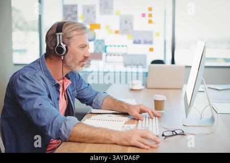 Homme d'âge moyen tapant sur le clavier blanc dans le bureau par fenêtre, avec casque, tasse à café, espace de copie Banque D'Images