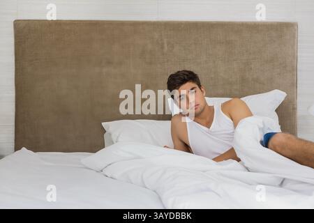 Homme incliné sur le lit dans la chambre, avec literie blanche grondée et tête de lit beige, espace copie Banque D'Images