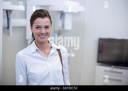 Femme debout dans la salle d'examen dentaire avec la machine à rayons X et le moniteur sur l'armoire blanche, souriant Banque D'Images