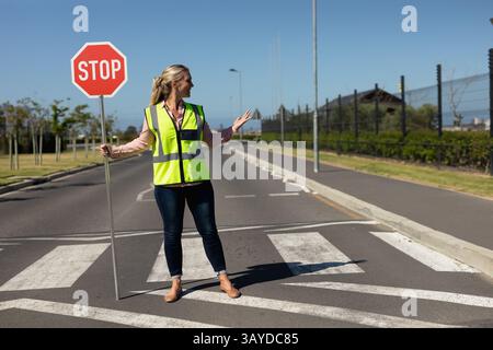 Femme tenant un panneau STOP rouge sur le passage à zèbre de banlieue, faisant des gestes près des bandes blanches du passage Banque D'Images