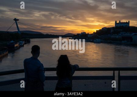 Un jeune couple observe le coucher du soleil sur le Danube dans la capitale slovaque Bratislava. En arrière-plan se trouvent la tour OVNI, le pont SNP, le château de Bratislava et la cathédrale de Martin. Banque D'Images