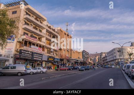 Scène de rue animée du moyen-Orient à Aqaba, en Jordanie : commerce occupé, signalisation arabe, voitures garées et piétons sous un ciel dégagé. Banque D'Images