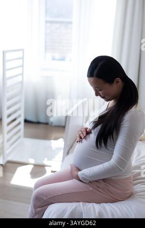 Femme enceinte asiatique assise sur le bord du lit dans la pépinière, berçant le ventre près de la crèche blanche, espace de copie Banque D'Images