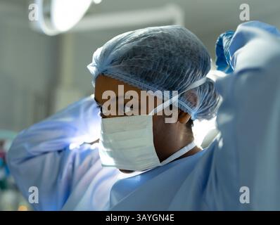 Chirurgien femme ajustant les sangles du masque et la casquette chirurgicale dans la salle d'opération, avec un plafonnier lumineux Banque D'Images