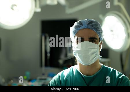Chirurgien masculin debout face à la caméra dans la salle d'opération, portant un masque chirurgical et une casquette sous les lumières Banque D'Images