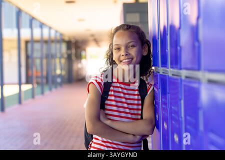 Fille multiraciale appuyée contre des casiers bleus dans le couloir de l'école, portant un sac à dos noir Banque D'Images