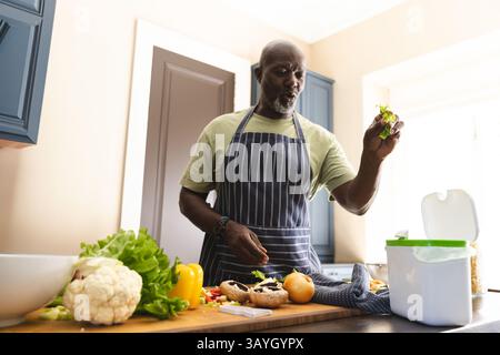 Homme afro-américain senior hachant des légumes dans la cuisine à la maison, avec bol à mélanger, bac à compost Banque D'Images
