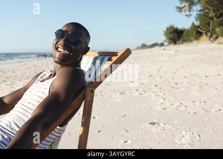 Homme afro-américain s'inclinant dans une chaise longue sur la plage, portant un dessus rayé, des lunettes de soleil, un espace de copie Banque D'Images