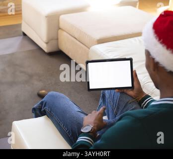 Homme afro-américain portant chapeau de Père Noël tenant tablette avec écran blanc dans le salon, espace de copie Banque D'Images