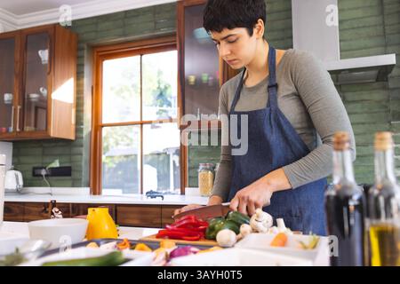 Jeune femme blanche hachant des légumes sur une planche à découper dans la cuisine de la maison, avec des pots et des bouteilles d'huile Banque D'Images