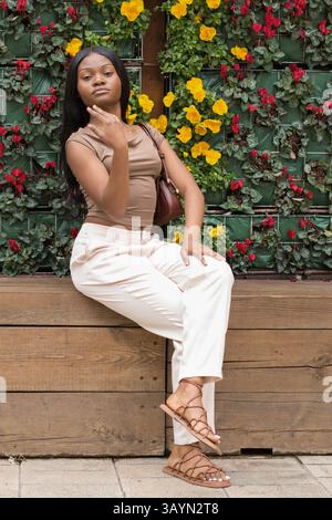 Jeune femme noire aux cheveux longs, portant un top de souris moka et un pantalon blanc, assise sur un banc en bois devant un mur floral, posant dans un s tendance Banque D'Images