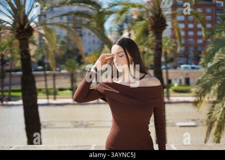 Jeune femme avec de longs cheveux bruns, portant une robe de couleur moka hors de l'épaule, posant dans un parc de la ville avec des palmiers et une pièce d'eau dans le Banque D'Images