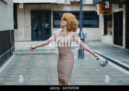 Jeune femme élégante avec des cheveux afro volumineux et des lunettes tient des écouteurs tout en profitant de la musique et en exprimant la joie dans un cadre urbain Banque D'Images