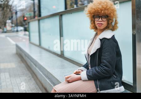 Jeune femme élégante avec des cheveux bouclés blonds et des lunettes est assise sur une marche en béton, portant des écouteurs et tenant un smartphone Banque D'Images