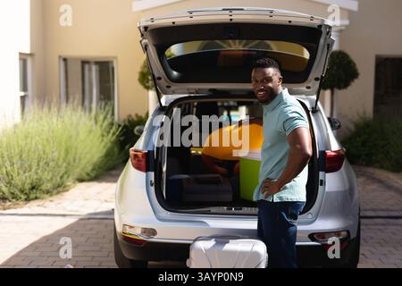 Homme afro-américain debout par coffre de SUV argenté dans l'allée, avec refroidisseur et valise, espace de copie Banque D'Images