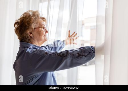 Femme âgée pairs espérons à travers de purs rideaux dans sa maison pendant la matinée. Humeur nostalgique, scène de confort à la maison pour le concept de style de vie de retraite a Banque D'Images