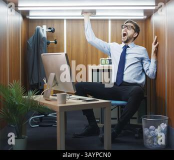 Employé fou désespéré pris au piège dans un minuscule bureau, il est assis à son bureau et crie Banque D'Images