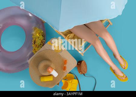 Jeune femme assise dans une chaise longue avec verre de cocktail et accessoires de plage sur fond bleu Banque D'Images