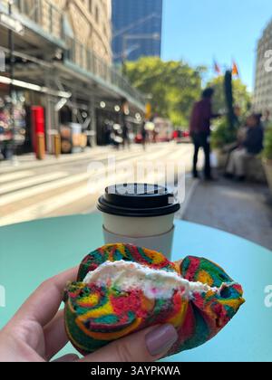 Main blanche féminine tenant bagel coloré et buvant du café contre le fond de la ville de New York le jour ensoleillé assis dans le café, photo verticale. Bonjour Banque D'Images