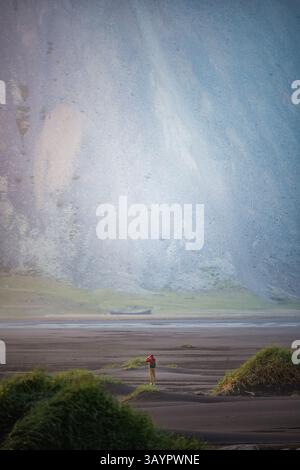 Seul randonneur avec veste rouge marche à travers les dunes de sable noir devant les falaises imposantes de la montagne Vestrahorn en Islande, évoquant la solitude. Banque D'Images