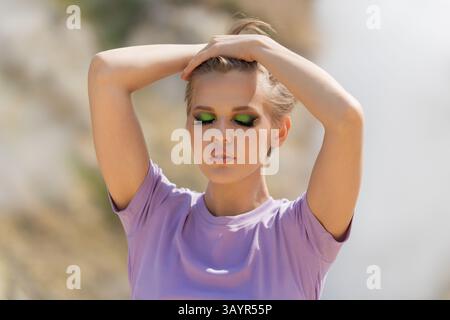 Portrait d'été de mannequin de mode a maquillage et leurs yeux sont fermés. Elle a les mains levées dans les cheveux. Femme pose sur fond naturel dans vo Banque D'Images