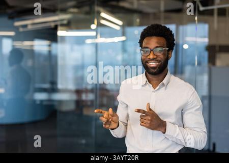 Un homme sympathique et souriant portant des lunettes pointe vers le côté dans un cadre de bureau moderne, mettant en valeur sa positivité. Banque D'Images