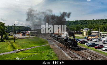 Une locomotive à vapeur longe les voies ferrées, dégageant des nuages de fumée sur fond de paysages verdoyants et d'une ancienne gare. Le paramètre Banque D'Images