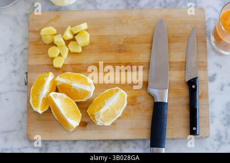 La planche à découper en bois tient des coins orange et du gingembre coupé en dés sur le comptoir en marbre près des couteaux, du verre Banque D'Images