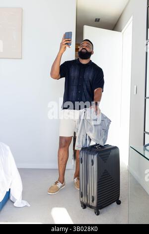 Homme afro-américain debout tenant smartphone par porte ouverte dans la chambre d'hôtel, avec valise Banque D'Images