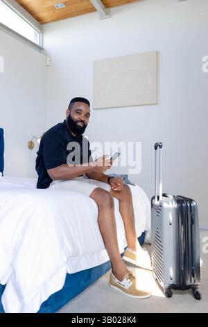 Assis homme afro-américain vérifiant smartphone sur le lit dans la chambre, avec valise roulante à proximité Banque D'Images