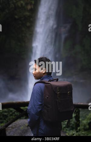 Homme asiatique en sweat à capuche bleu voyageant à une cascade avec sac à dos brun Banque D'Images