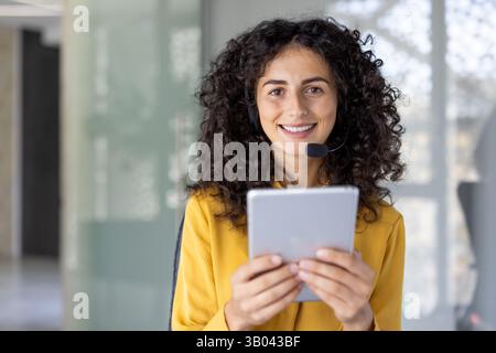 Une femme souriante portant un micro-casque tient une tablette, paraissant prête pour le service client dans un environnement de bureau moderne. Banque D'Images