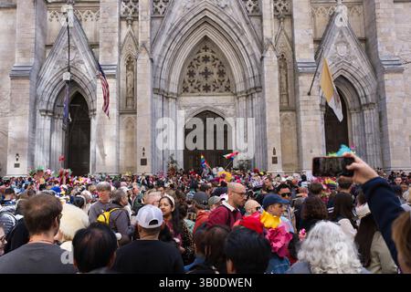 Une grande foule de gens, dont beaucoup portent des bonnets de Pâques, se rassemble devant une grande cathédrale de pierre. Les drapeaux américain et du Vatican sont visibles. Une personne u Banque D'Images