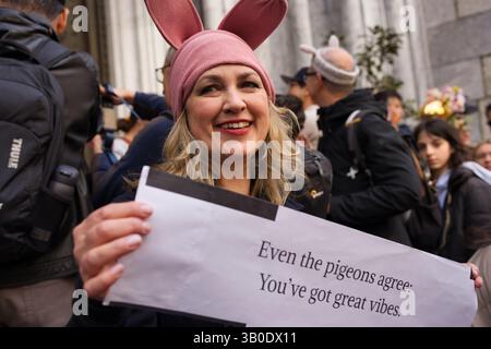 Une femme à la peau claire avec les cheveux blonds, portant un chapeau d'oreille de lapin rose, tient un panneau qui dit, "même les pigeons sont d'accord : vous avez de grandes vibrations." Elle i Banque D'Images