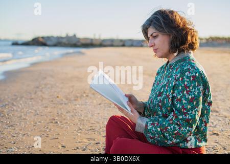 Femme d'âge moyen se relaxant sur la plage au coucher du soleil, immergée dans un livre tout en étant assise sur le sable chaud Banque D'Images