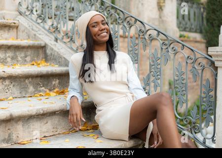 Jeune femme noire souriante assise sur les escaliers avec des feuilles d'automne dans un parc Banque D'Images