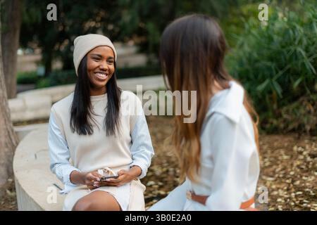 Deux amies sont assises dans un parc, tenant leur smartphone et ayant une conversation, profitant d'un moment de détente et de connexion au milieu Banque D'Images