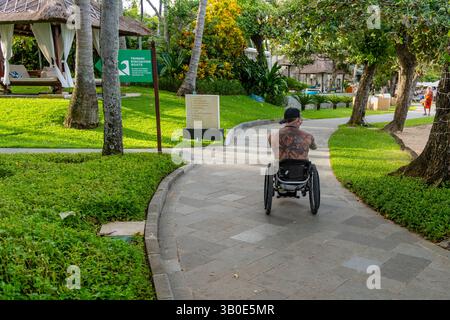 Bali, Indonésie - 19 décembre 2024 : un homme en fauteuil roulant roule sur un chemin dans un parc. Le chemin est bordé d'arbres et d'herbe, et il y a un panneau à proximité Banque D'Images